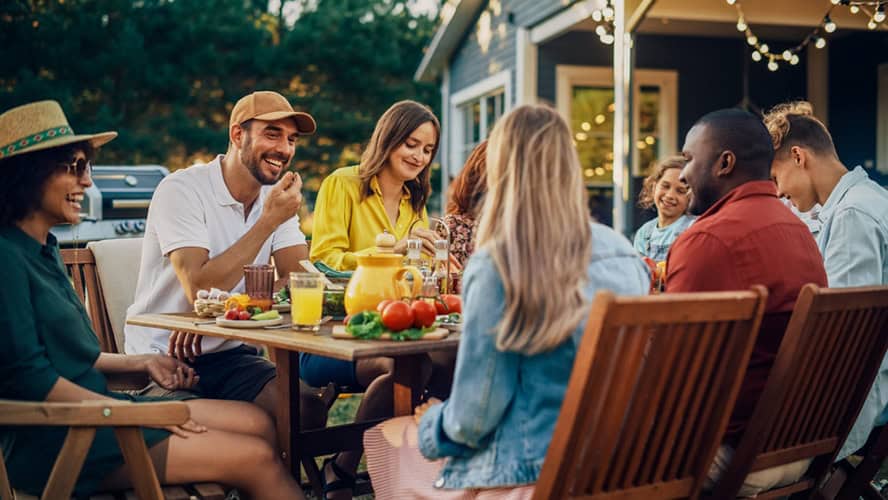 A group of people sit around a wooden outdoor table, enjoying a meal together under the twinkling string lights. The backyard setting, with its cheerful ambiance and pest control in Suffolk County NY ensuring a comfortable evening, displays a spread of food and a charming yellow teapot.