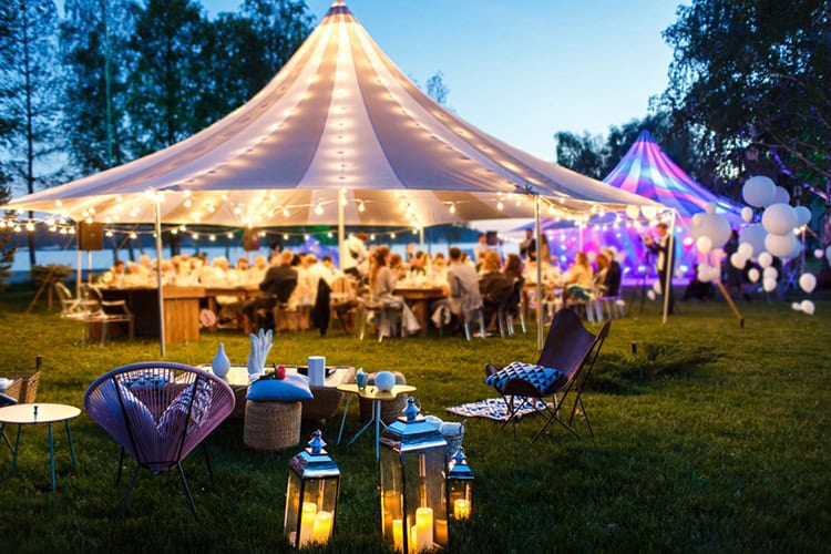 A large outdoor tent, illuminated with string lights, hosts a gathering. People are seated at tables, while lanterns and chairs adorn the grass. The festive scene at dusk benefits from expert pest control in Suffolk County, NY, ensuring a comfortable atmosphere for all.