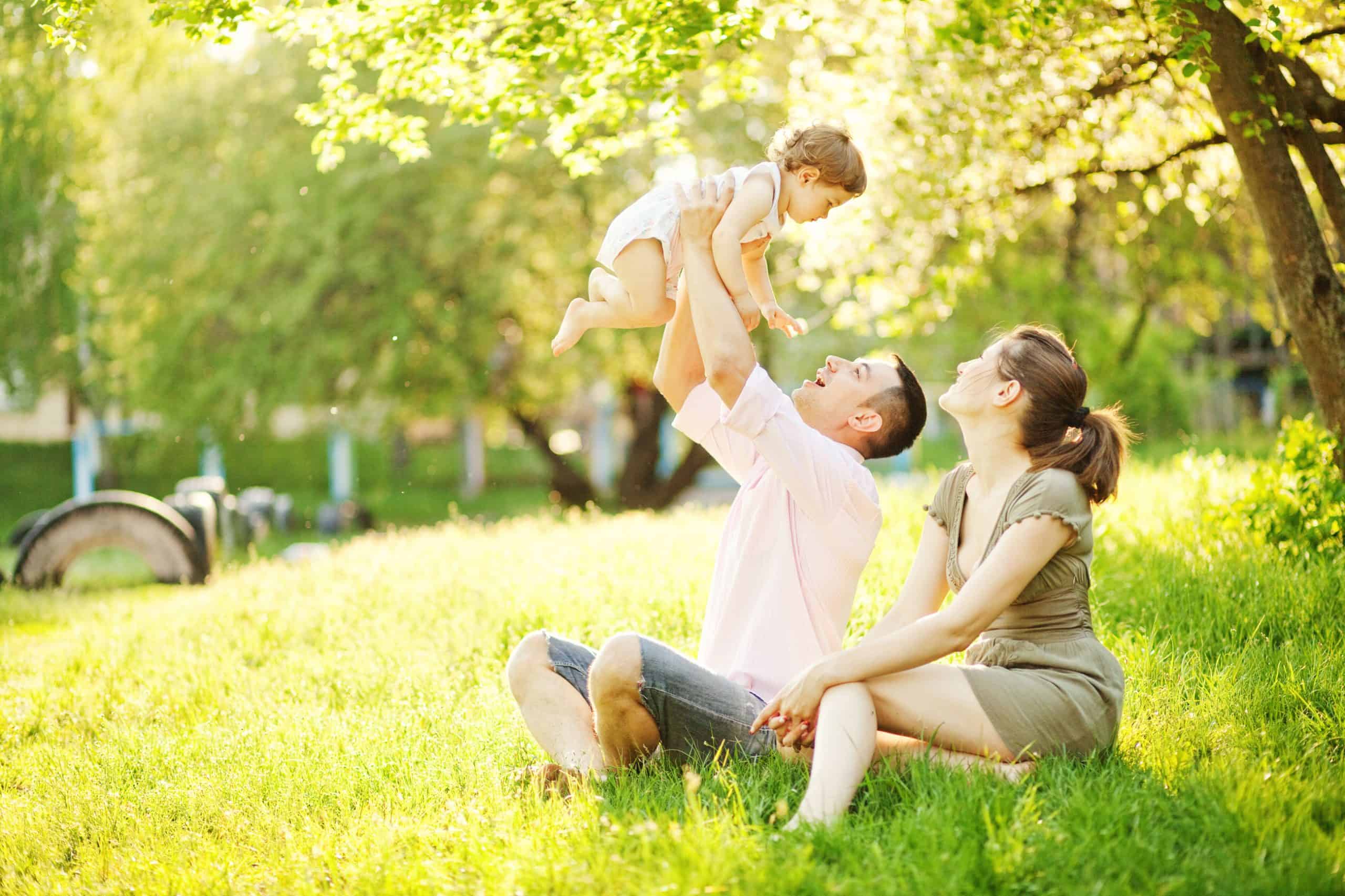 A man and woman sit on grass in a sunlit park, enjoying the warm day. The man, dressed in a light shirt and shorts, joyfully lifts a smiling baby up into the air. The woman in a green dress watches with delight, mindful of tick control in Suffolk County NY. Trees and a stroller rest peacefully in the background.