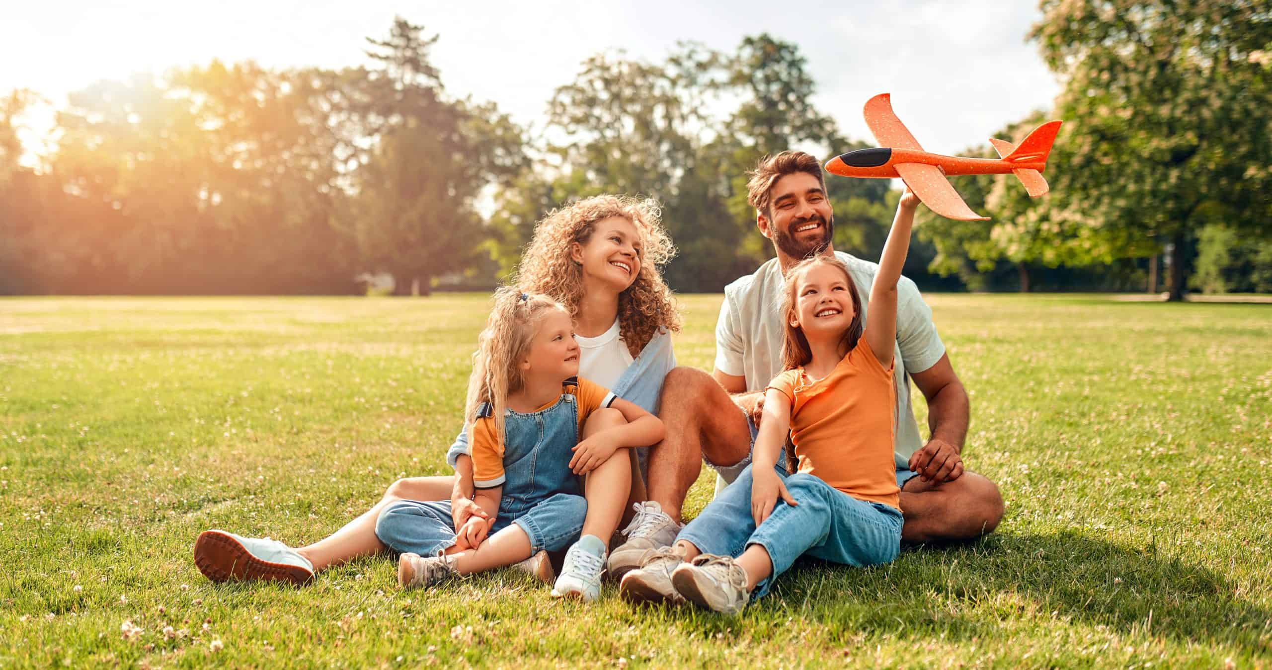 A family of four enjoys a sunny park in Suffolk County, NY. A man and woman smile as two young girls play with a toy airplane, safe from ticks thanks to effective pest control. Trees stand tall in the background, with sunlight filtering through to create a warm, cheerful atmosphere.