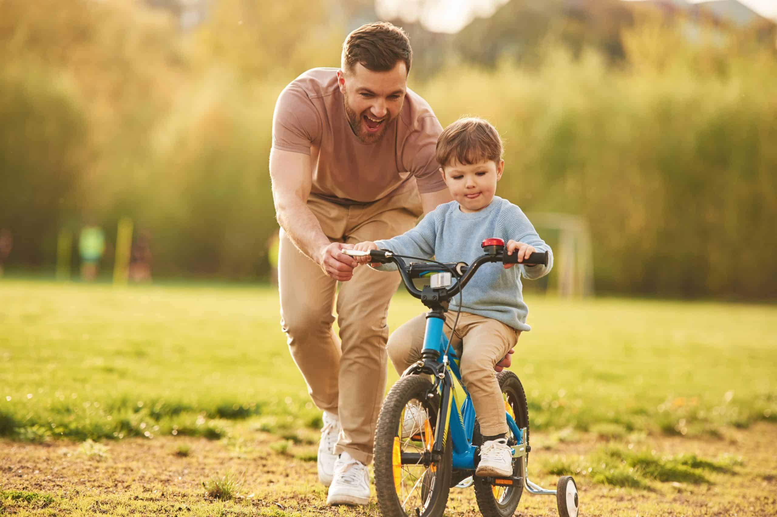 A man joyfully helps a young child ride a blue bicycle with training wheels on a grassy field recently treated for tick control in Suffolk County, NY. Both appear happy and engaged in the activity under the clear sky.