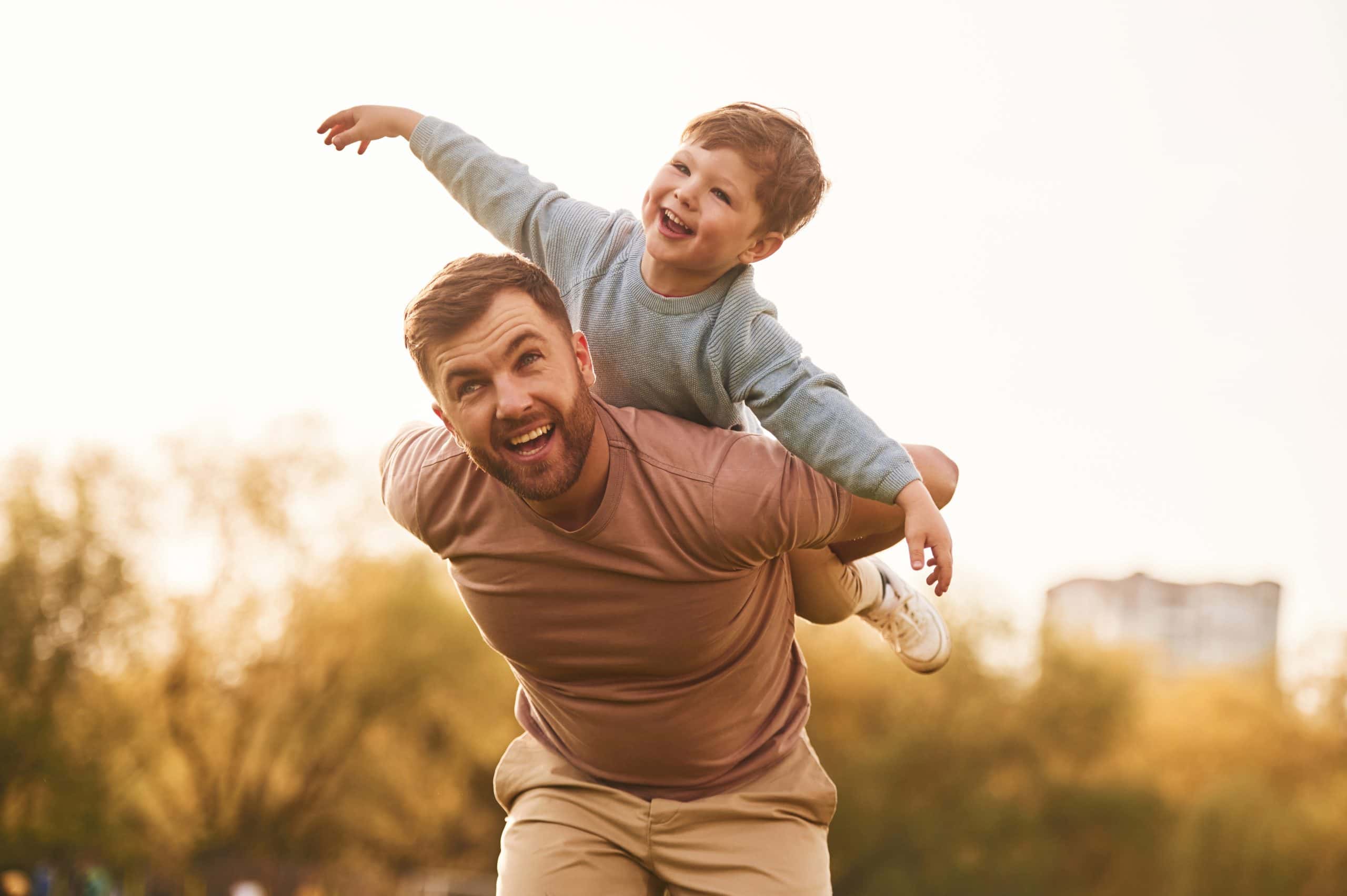 A man in a brown shirt carries a joyful child on his back, the boy spreading his arms out like wings. They revel outdoors amid trees and a building under a clear sky, enjoying carefree moments thanks to effective pest control in Suffolk County, NY.