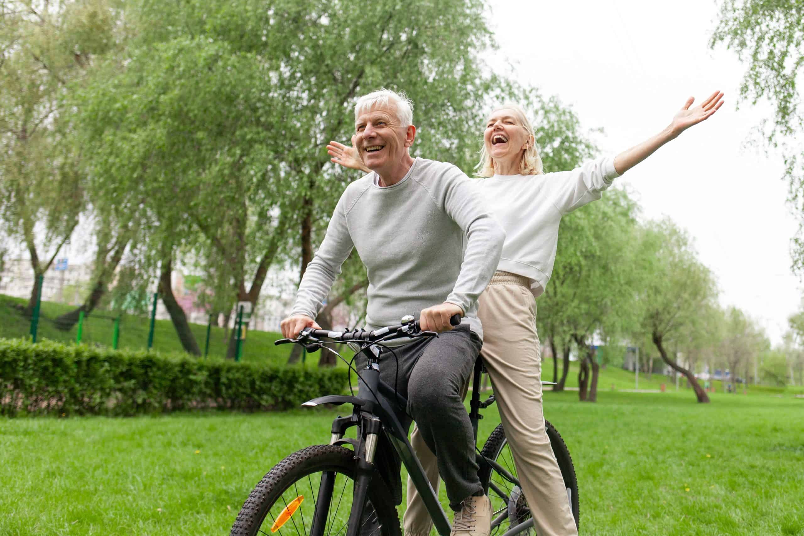 An older couple joyfully rides a bicycle together in a serene Suffolk County, NY park. The man pedals while smiling, and the woman sits with arms outstretched, laughing amidst lush green grass and trees—untouched by worry thanks to effective tick control.