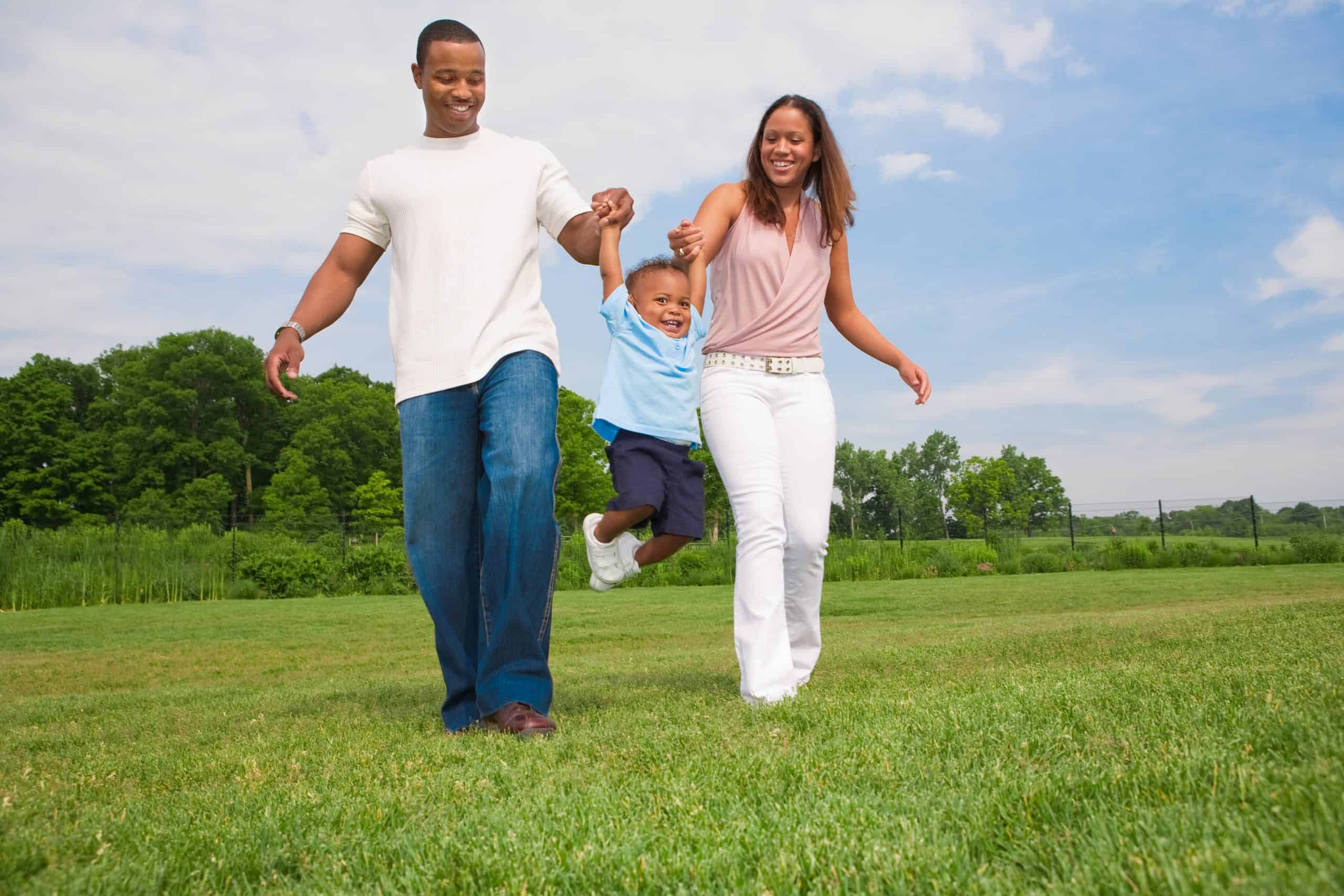 A family of three enjoys a sunny day outdoors on a field in Suffolk County, NY. The father and mother hold hands with their young child, swinging him playfully as they savor tick-free peace of mind thanks to expert tick control services. Trees and a partly cloudy sky frame the joyful scene.